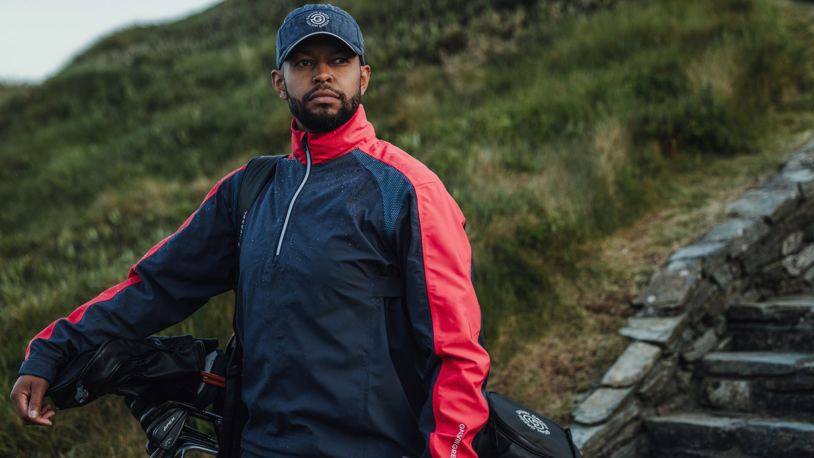A male golfer carrying his bag, wearing a rain jacket and rain cap