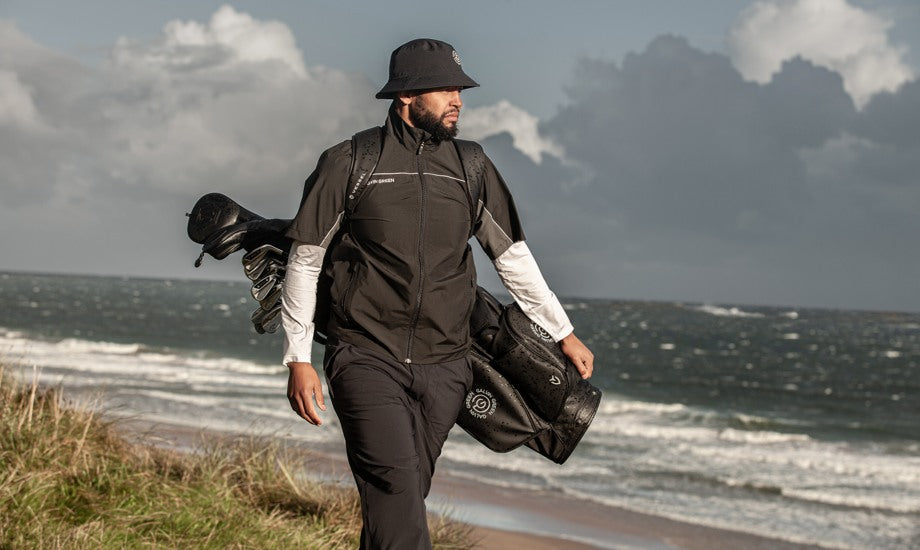 A male golfer walking next to water on a golf course, wearing black and white outfit