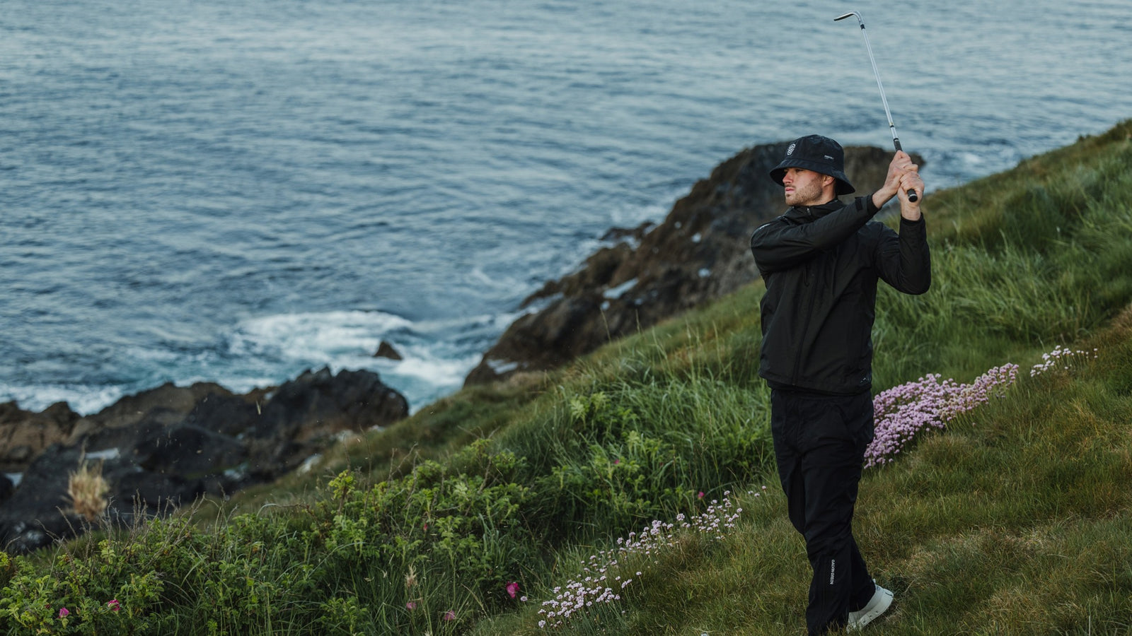 A male golfer swinging his club, wearing black waterproof golf gear.