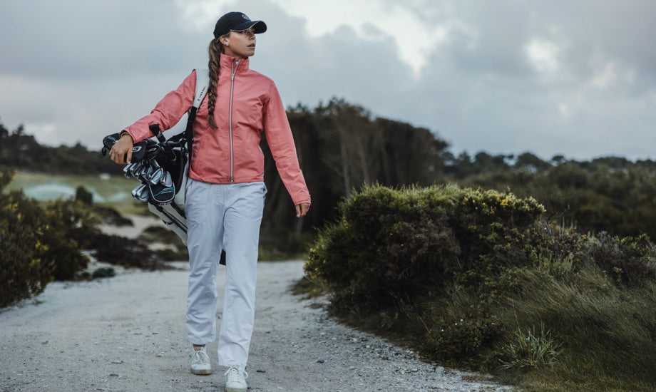 A female golfer walks through a golf course wearing waterproof cute golf clothes and carrying her clubs.