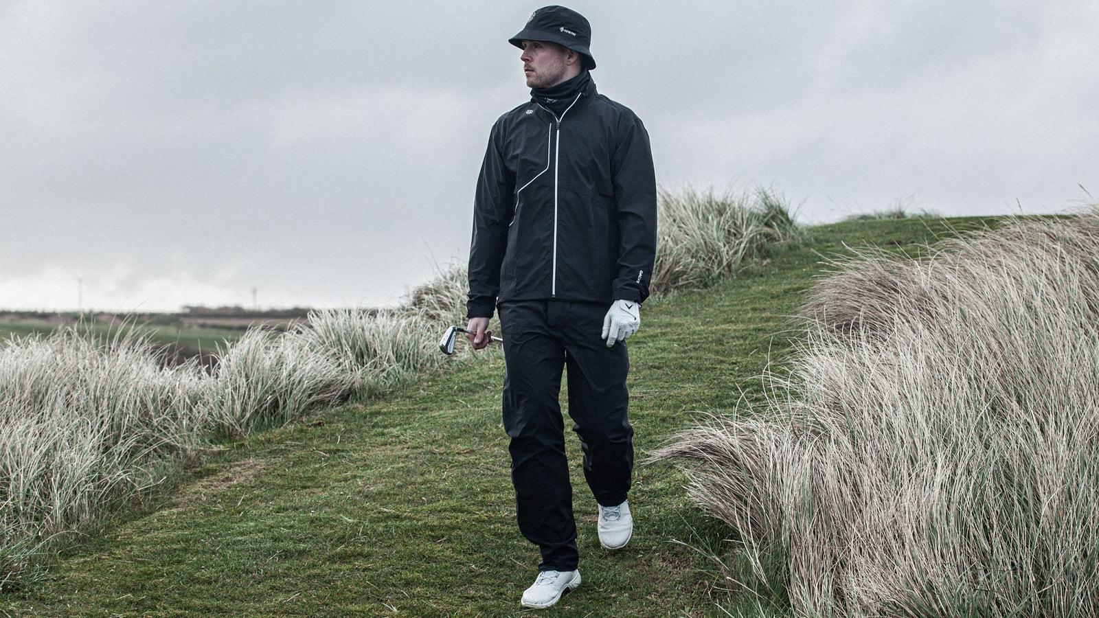 A male golfer walking on a golf course holding a golf club and wearing a black waterproof outfit.