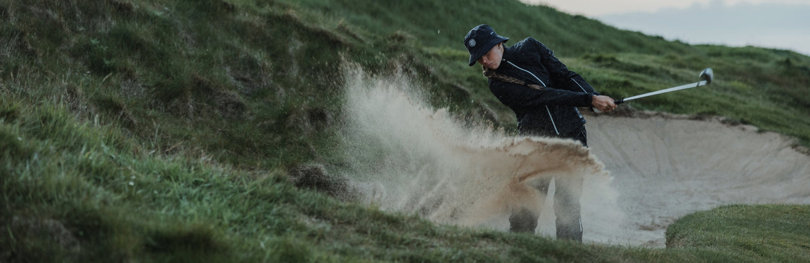 A women is doing a bunker shot and the sand is splashing. She is wearing golf rain gear in black