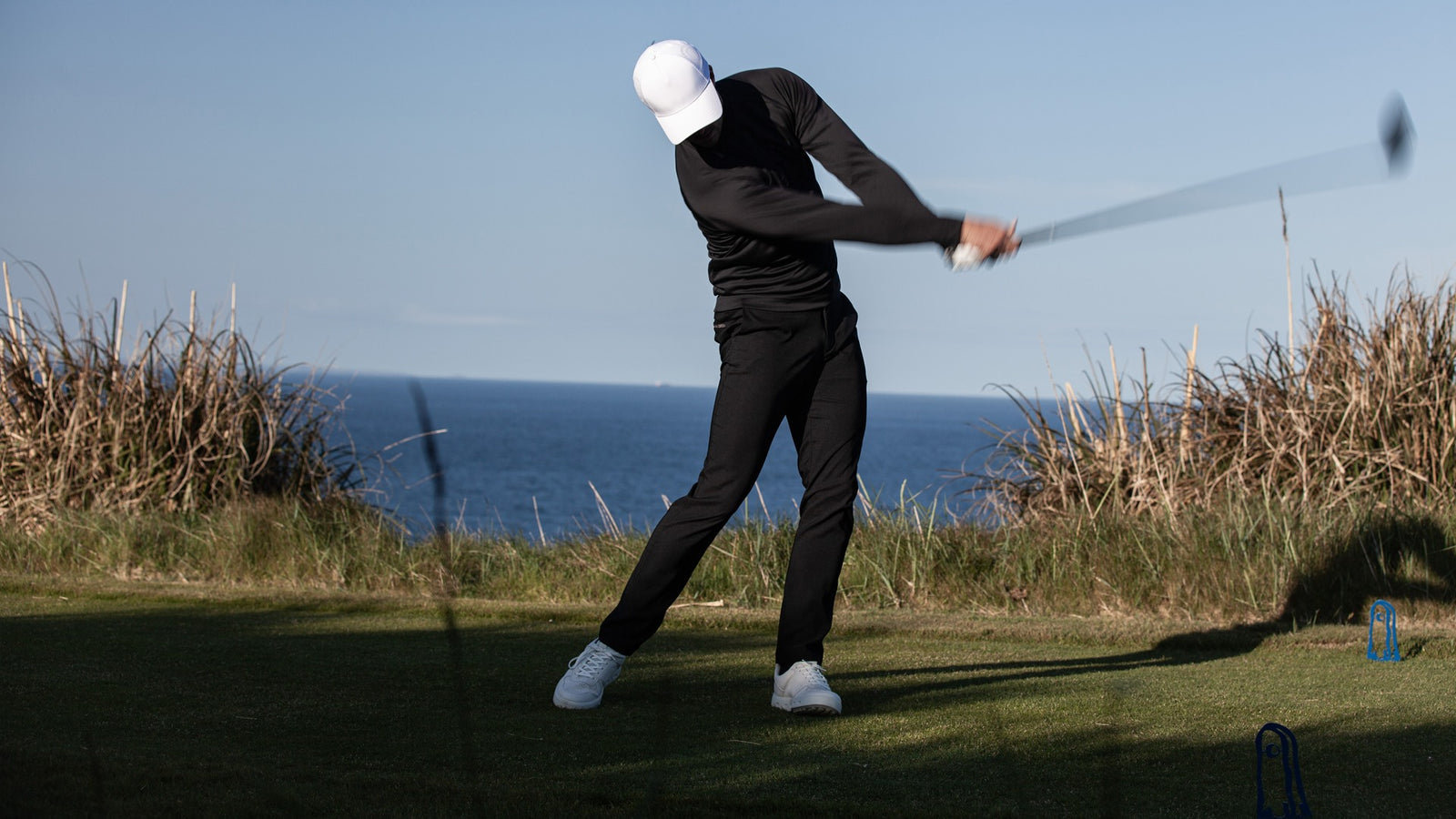 A golfer swinging his club wearing black clothes and a white cap