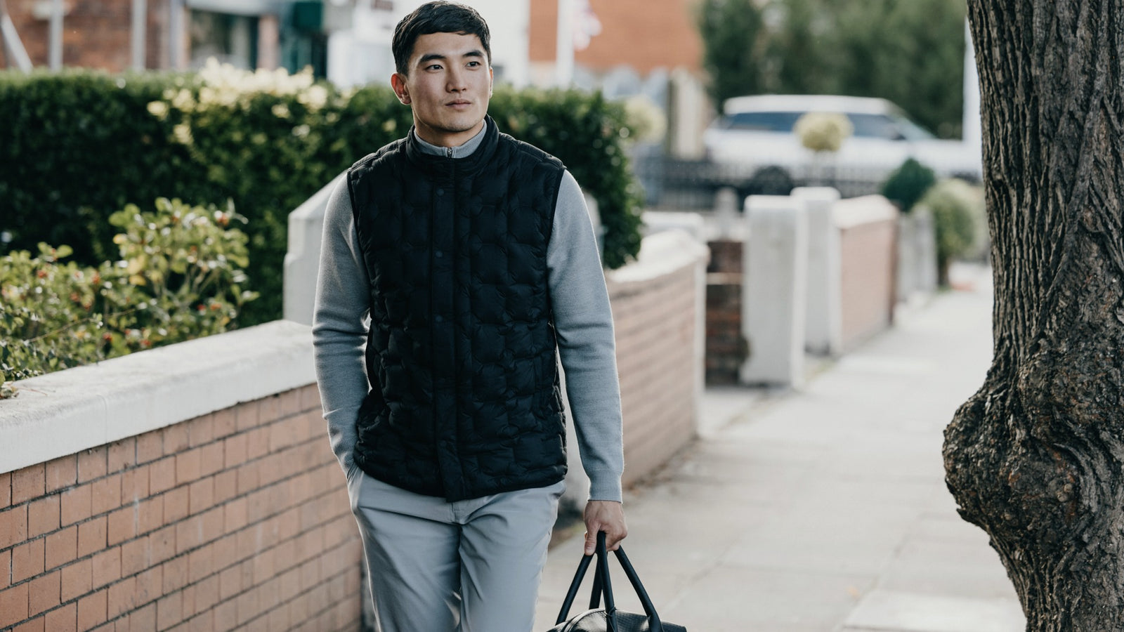 A male walking on a sidewalk holding a bag wearing grey clothes with a black vest